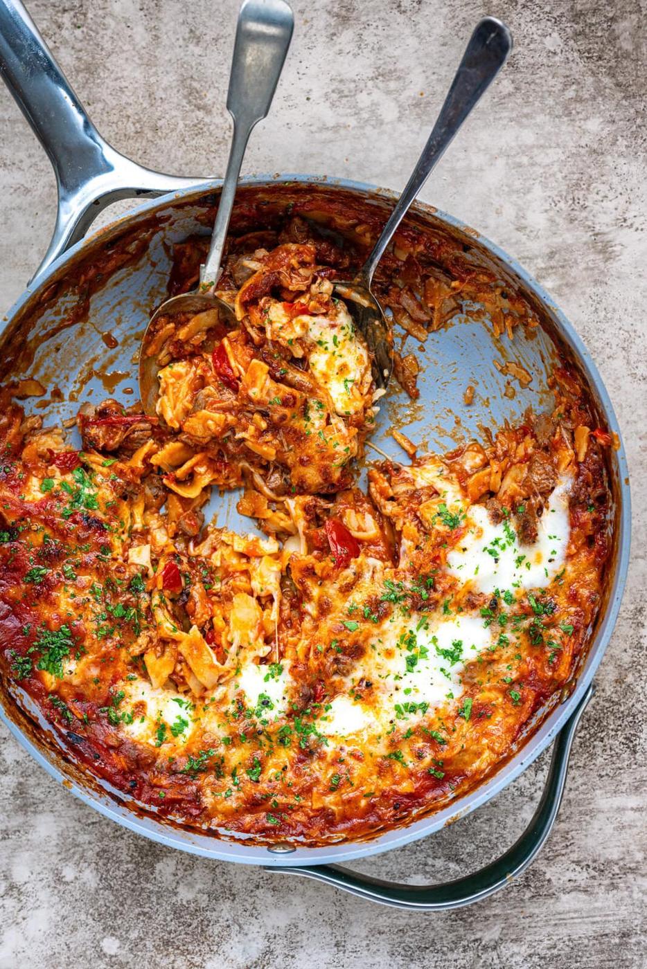 A skillet filled with cheesy lasagne topped with parsley. Two serving utensils rest in the pan, with a portion of the pasta scooped out, revealing a saucy, golden, and slightly browned surface.