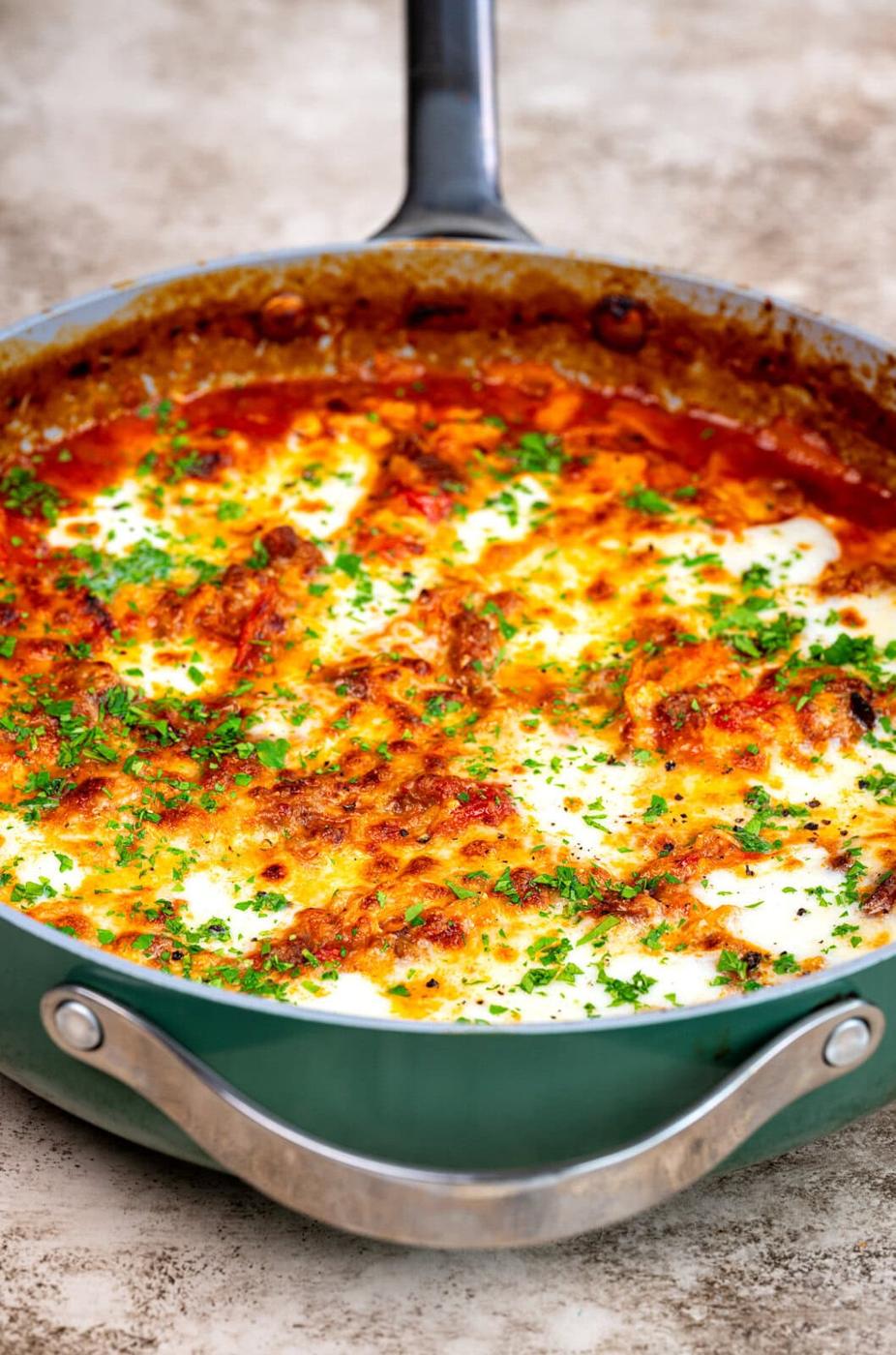 A skillet filled with baked lasagna, topped with melted cheese, browned edges, tomato sauce, and sprinkled with fresh chopped parsley.