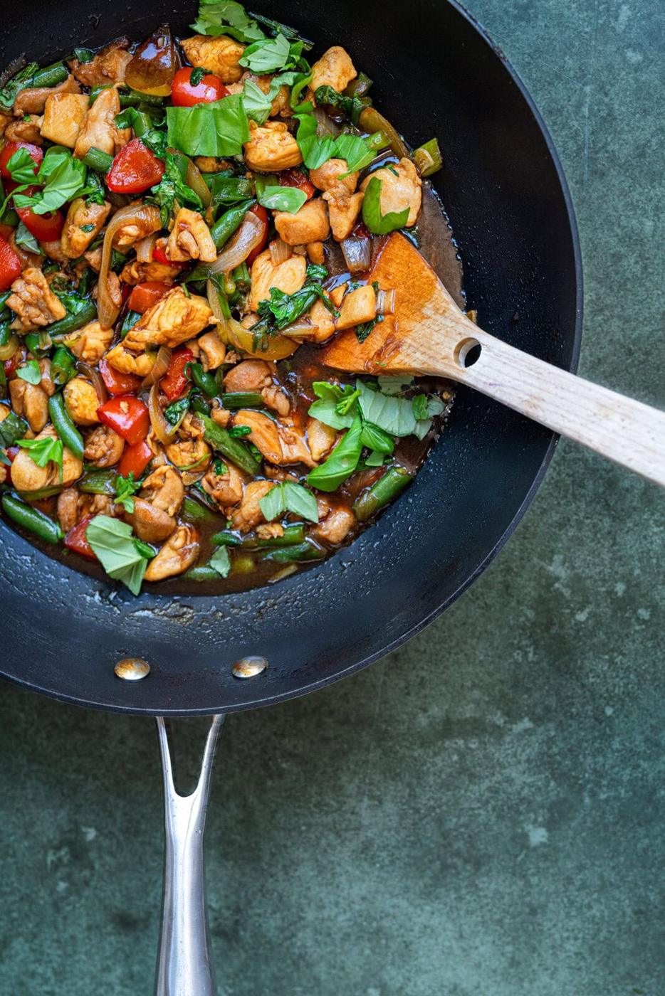 A black wok with Thai basil chicken, stir-fried vegetables, and fresh basil, with a wooden spoon resting inside, on a dark green countertop.