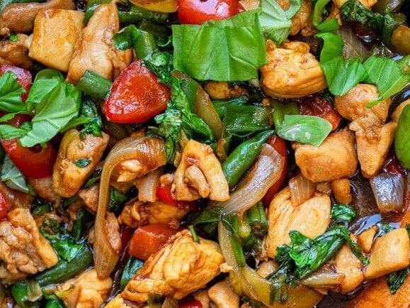 A black skillet filled with Thai basil chicken, stir-fried vegetables, and fresh basil, with a wooden spoon resting inside, on a dark green countertop.