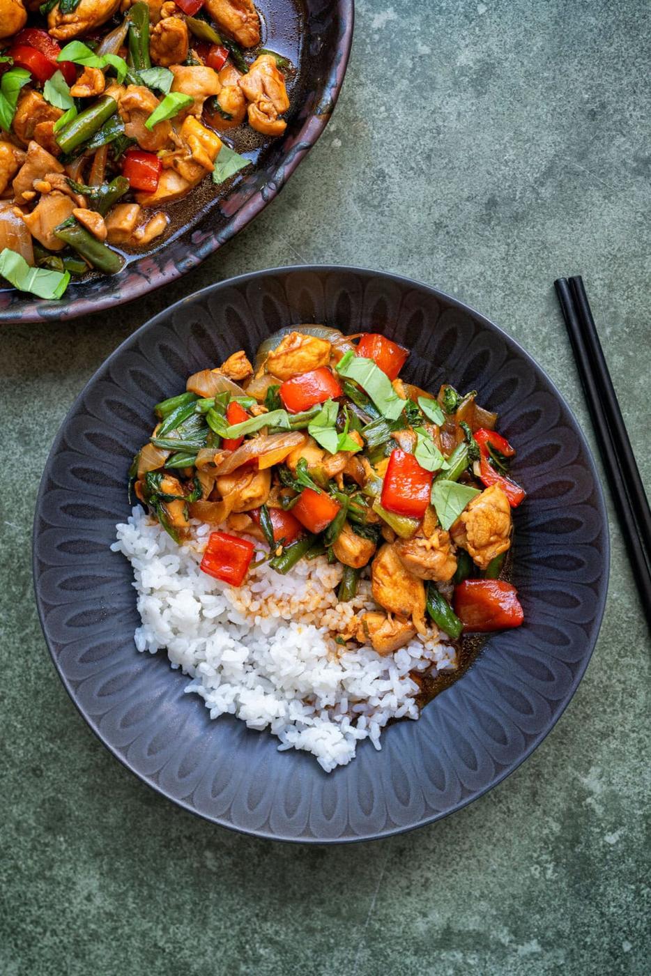 A black bowl with white rice topped with stir-fried Pad Krapow chicken, red bell peppers, green onions, and leafy greens, placed next to black chopsticks on a gray surface.