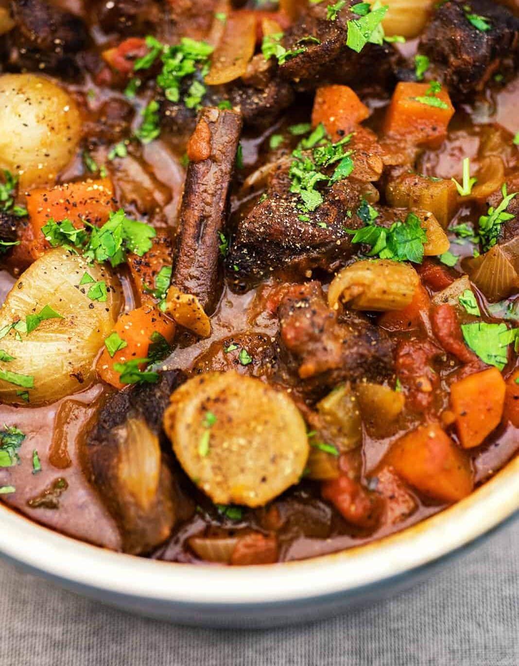 A close-up of a hearty Greek beef stew, or beef stifado, with chunks of beef, whole pearl onions, carrots, and herbs in a rich brown broth, garnished with chopped parsley.