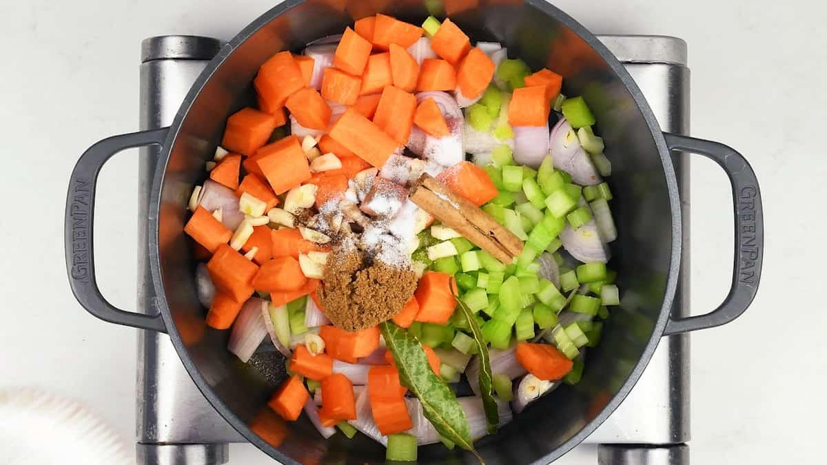 Chopped carrots, celery, onions, garlic, spices, a cinnamon stick, and a bay leaf are placed in a black pot on a stovetop—classic ingredients for Beef stifado, a traditional Greek beef stew, ready for cooking.