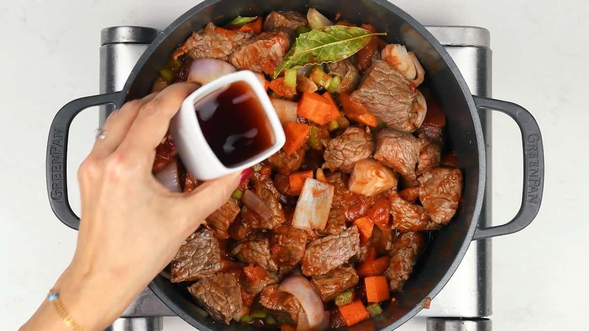 A hand pours a dark liquid from a small white cup into a pot filled with chunks of beef, carrots, onions, and bay leaves on a stovetop—perfect for preparing a classic beef stifado recipe.