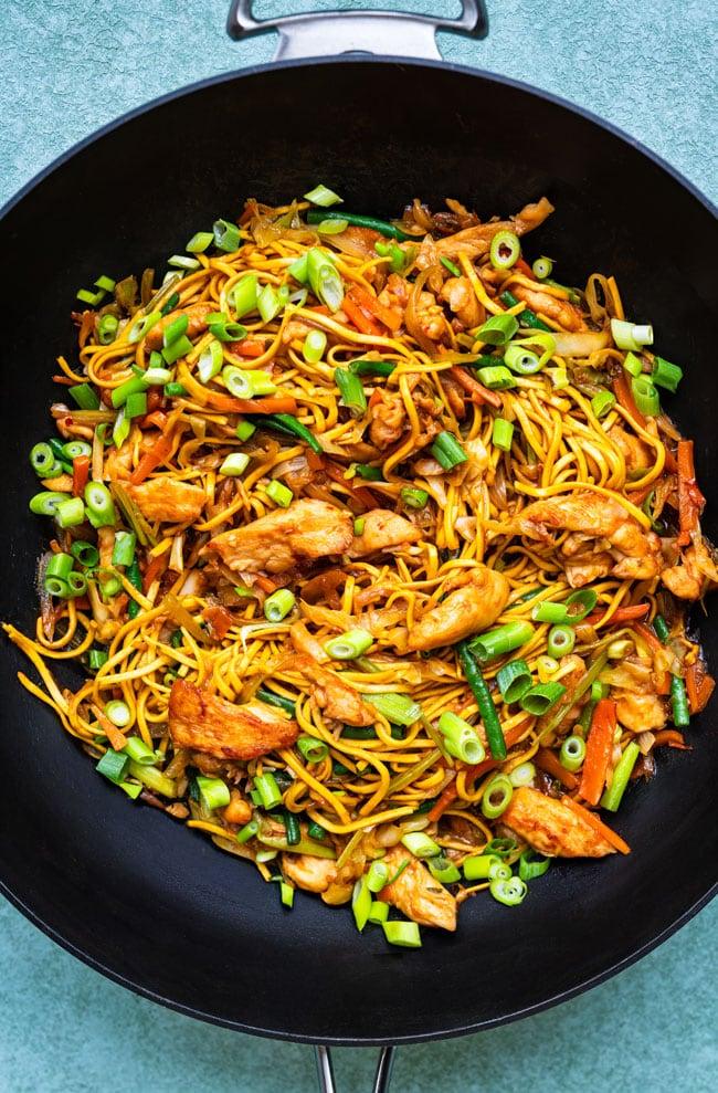 A wok filled with chicken chow mein - stir-fried noodles, sliced chicken, carrots, green onions, and other vegetables, garnished with chopped green onions on top.