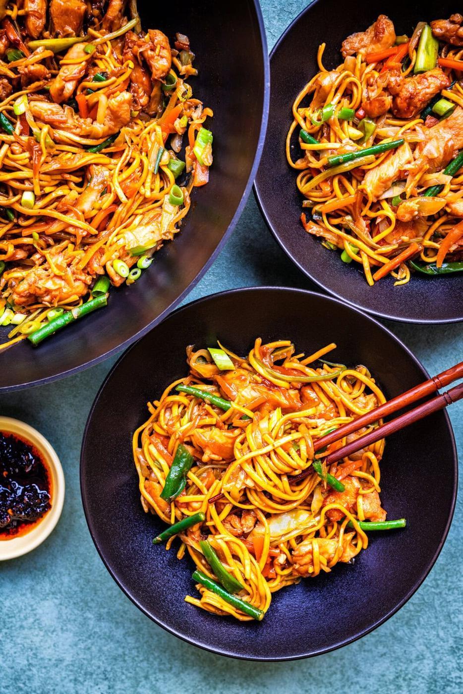 Three black bowls filled with chicken chow mein: noodles, chicken, green beans, and sliced vegetables. A pair of chopsticks rests on one bowl, and a small dish of red chili sauce sits nearby on a blue surface.