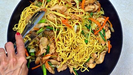 A hand using tongs to mix chow mein noodles, sliced chicken, and assorted vegetables—carrots, green beans, and onions—in a large black pan.