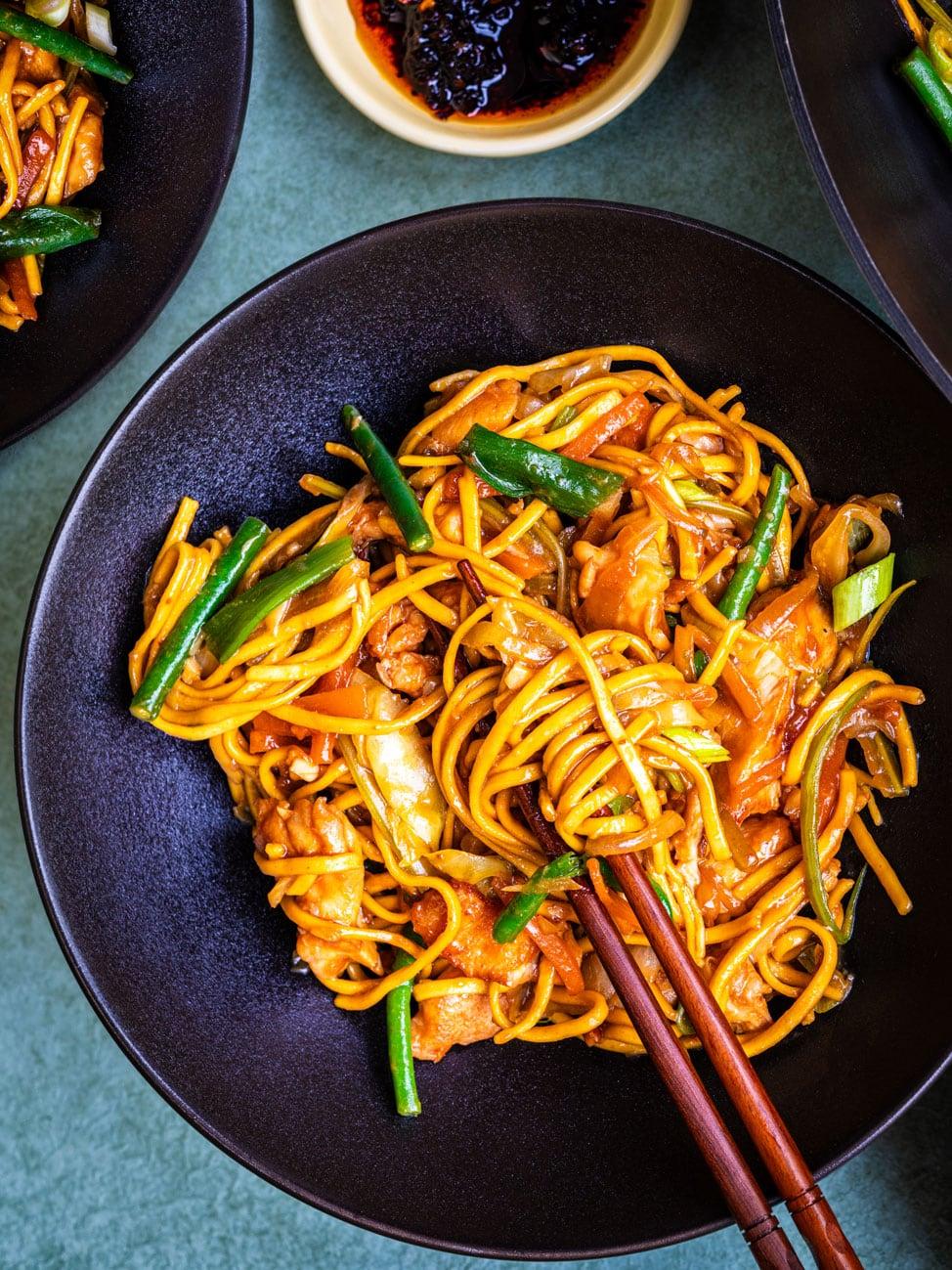 A bowl of stir-fried noodles with vegetables and chicken, garnished with green onions, is being picked up with chopsticks. A small dish of chili sauce is visible in the background.