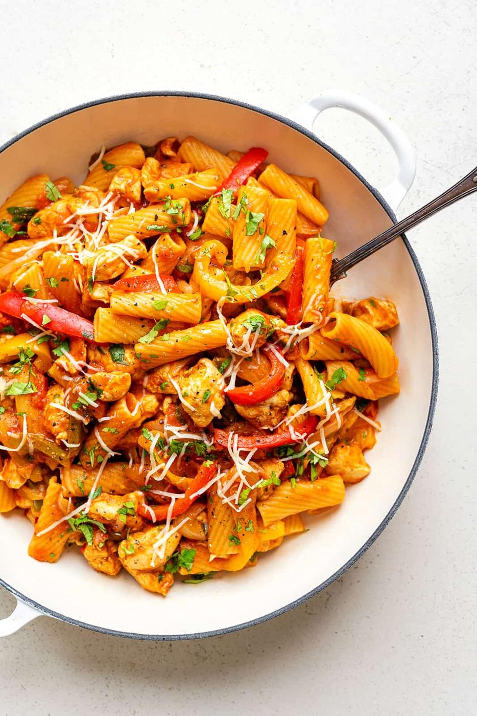 A white pan filled with One-Pot Chicken Fajita Pasta—rigatoni, cooked chicken, red bell peppers, tomato sauce, and topped with shredded cheese and parsley. A fork rests inside the pan.
