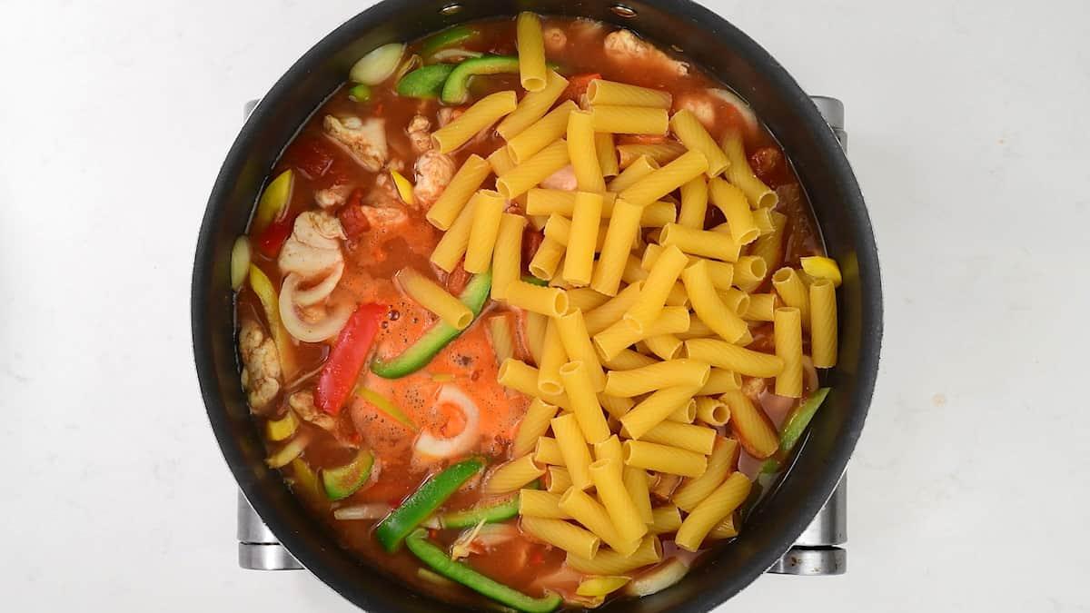 A pot of One-Pot Chicken Fajita Pasta with raw penne, green bell peppers, onions, tomatoes, and chicken chunks in a red broth on a white surface.