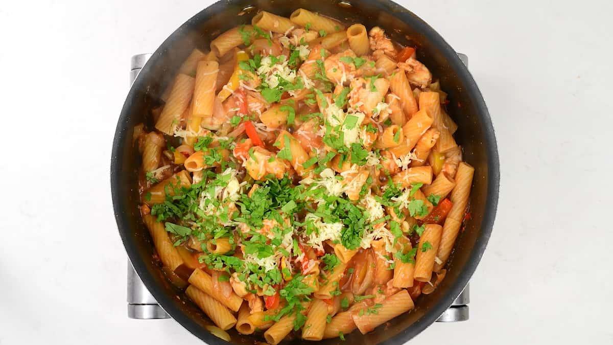 A pot of cooked rigatoni pasta mixed with tomato sauce, vegetables, and tender chicken, inspired by One-Pot Chicken Fajita Pasta, topped with grated cheese and fresh chopped parsley, seen from above on a white surface.