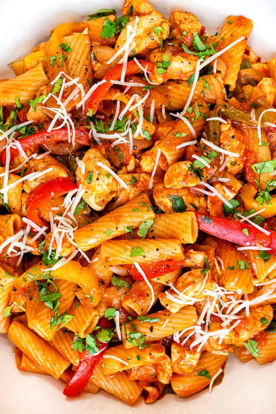 A close-up of One-Pot Chicken Fajita Pasta features rigatoni mixed with chicken, red bell peppers, herbs, and topped with shredded parmesan cheese.