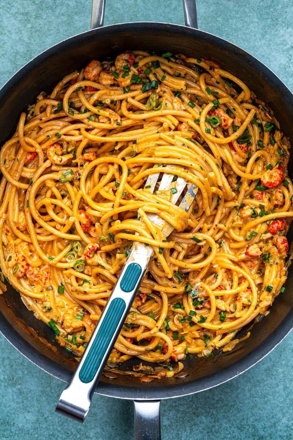 A pan filled with creamy crawfish pasta cooked in one saucepan, mixed with small pieces of seafood and garnished with chopped herbs. A slotted spoon is resting in the pasta, ready to serve.