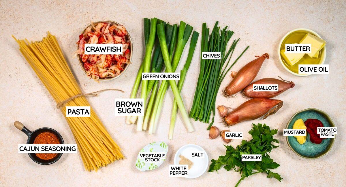 Top-down view of ingredients for a dish, including labelled items: pasta, crayfish, spring onions, brown sugar, chives, shallots, butter, olive oil, Cajun seasoning, vegetable stock, white pepper, salt, garlic, parsley, mustard and tomato purée.