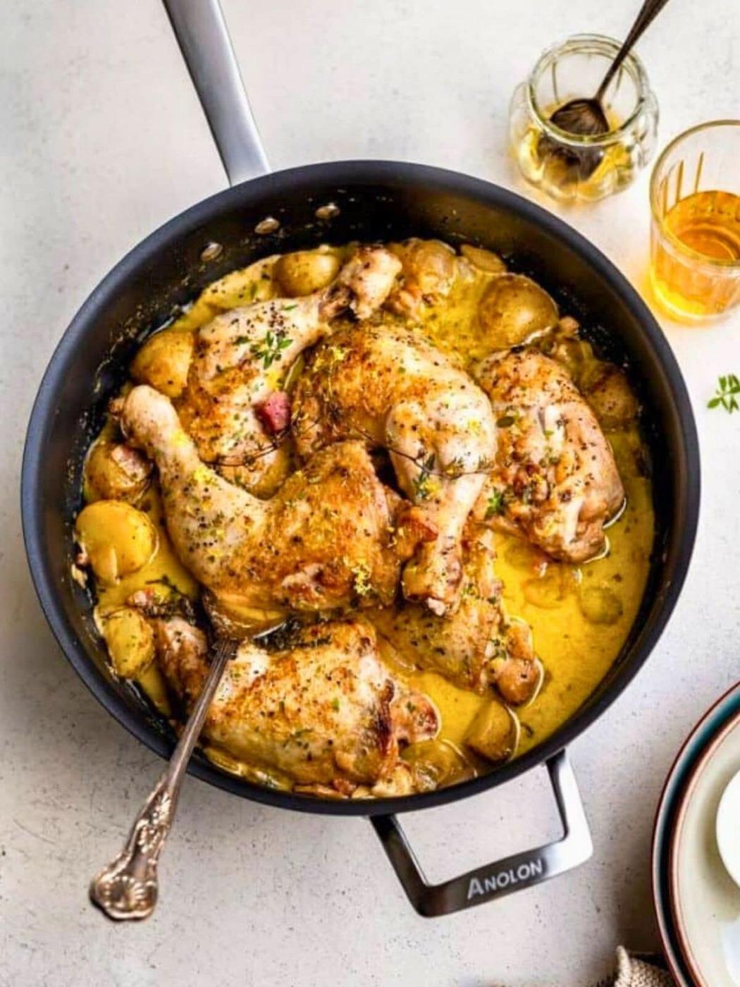 A pan of creamy chicken casserole featuring cooked drumsticks and potatoes in a rich yellow sauce, garnished with herbs, sits on a light countertop next to a drink, a jar with a spoon, and stacked plates with a napkin.