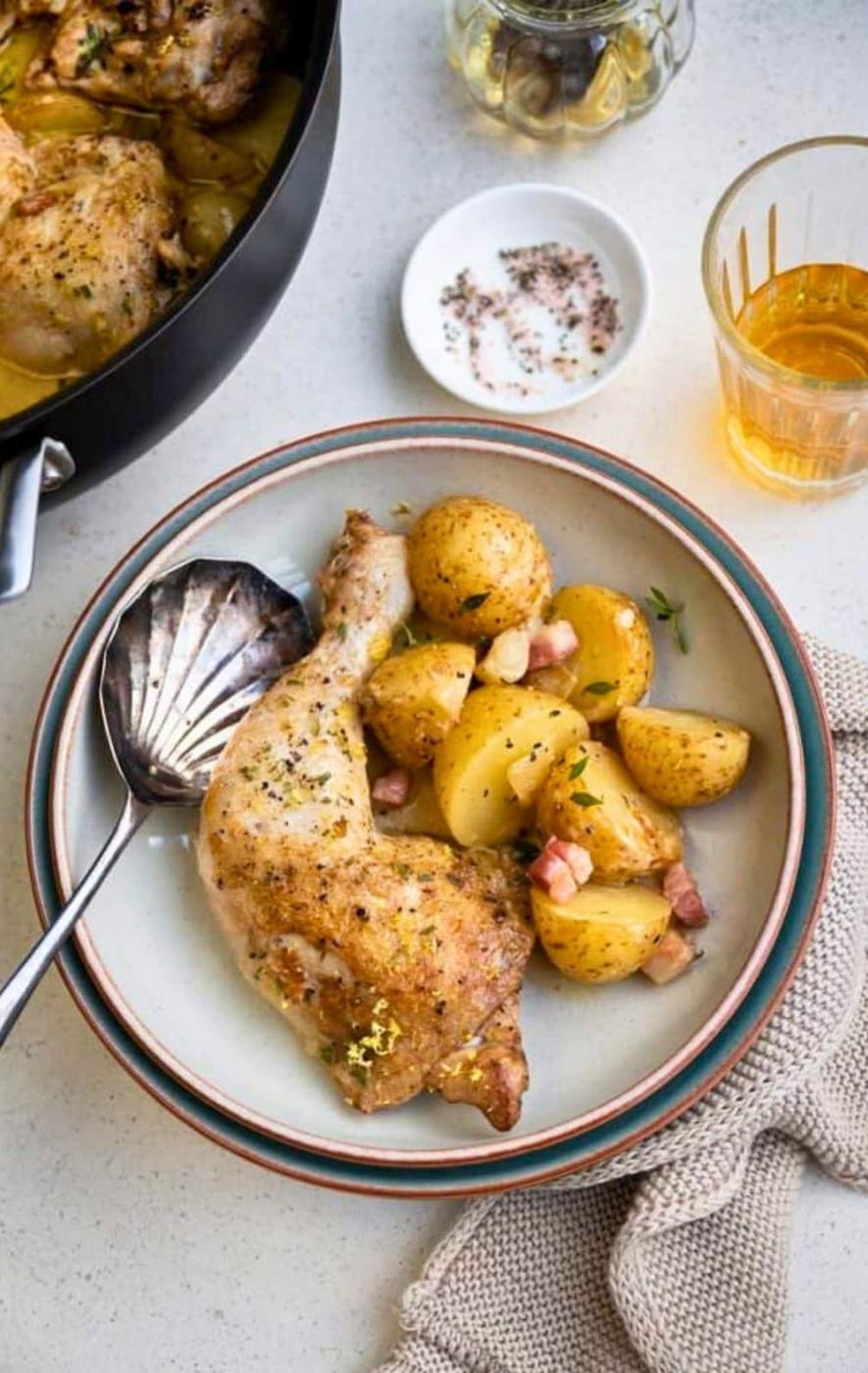 A plate with a roasted chicken leg, sliced golden potatoes, and small bacon pieces, garnished with herbs—reminiscent of a creamy chicken casserole. A serving spoon, a glass of amber drink, seasoning bowl, and textured cloth sit nearby.