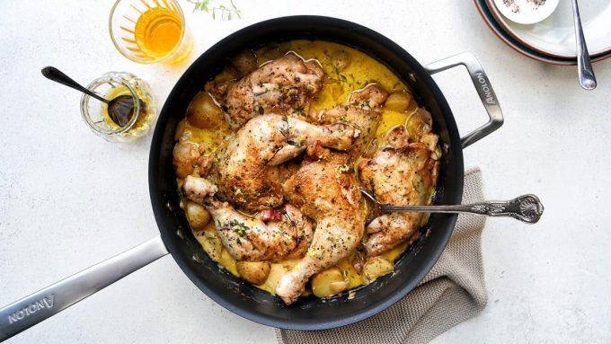A creamy chicken casserole pan with roasted chicken legs, small potatoes, and a rich yellow sauce, garnished with herbs. A spoon rests in the pan, next to a glass of drink and a small jar on a light-colored table.