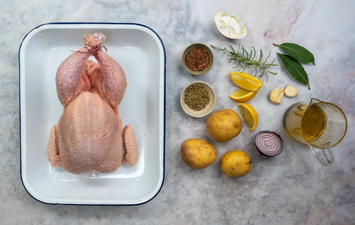 Ingredients for Greek roast chicken and lemon potatoes: a whole raw chicken, potatoes, red onion, lemon, garlic, dried oregano, bay leaves and olive oil on a marble surface