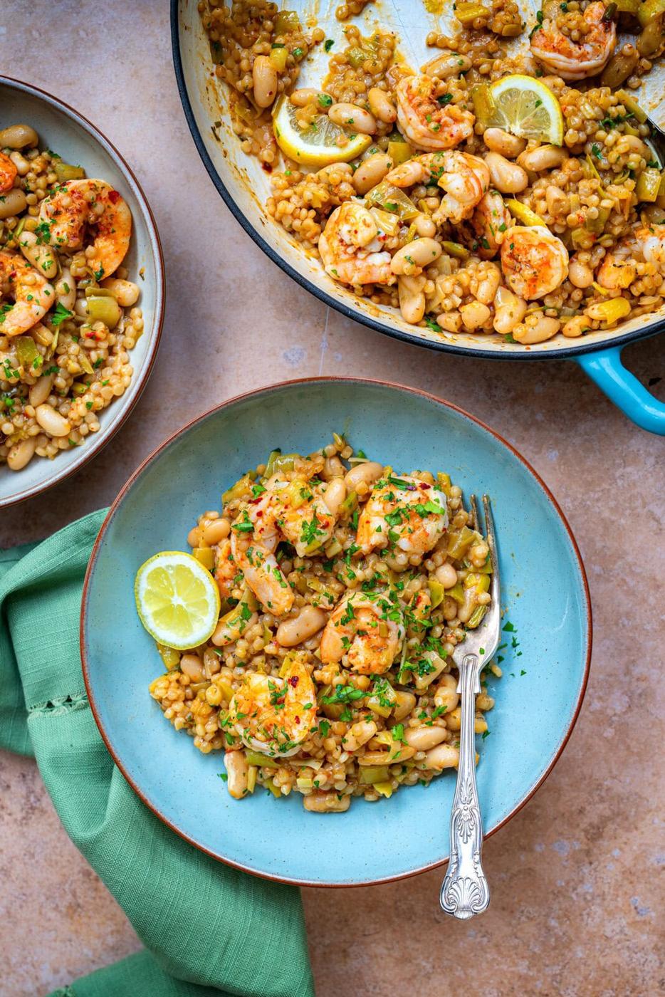 A bowl of shrimp with lemon, beans, and couscous garnished with herbs. A fork rests on the side of the bowl. A similar dish is served in a pan in the background. A green cloth napkin is beside the bowl.