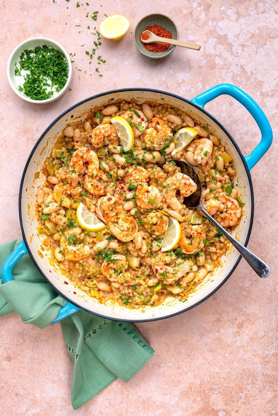 A colorful dish in a blue-handled pot features shrimp, white beans, leeks, giant couscous and herbs. Lemon slices are scattered on top. A green napkin lies nearby. Small bowls of chopped herbs, ground spice, and half a lemon are above.