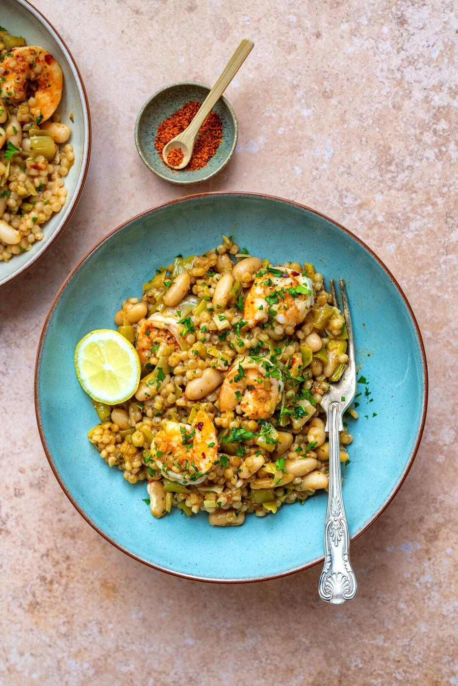 A blue bowl filled with shrimp, white beans, and pearl couscous, garnished with fresh herbs and a lime wedge. A fork rests on the bowls edge. A small bowl of red seasoning is placed in the background on a light-textured surface.