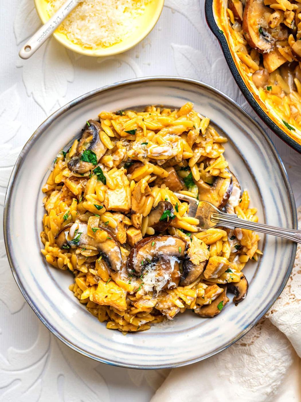 A plate of creamy orzo pasta mixed with mushrooms and herbs, topped with grated cheese. A fork rests on the plate, and a dish of cheese and a pan with more orzo are nearby on a white textured surface.