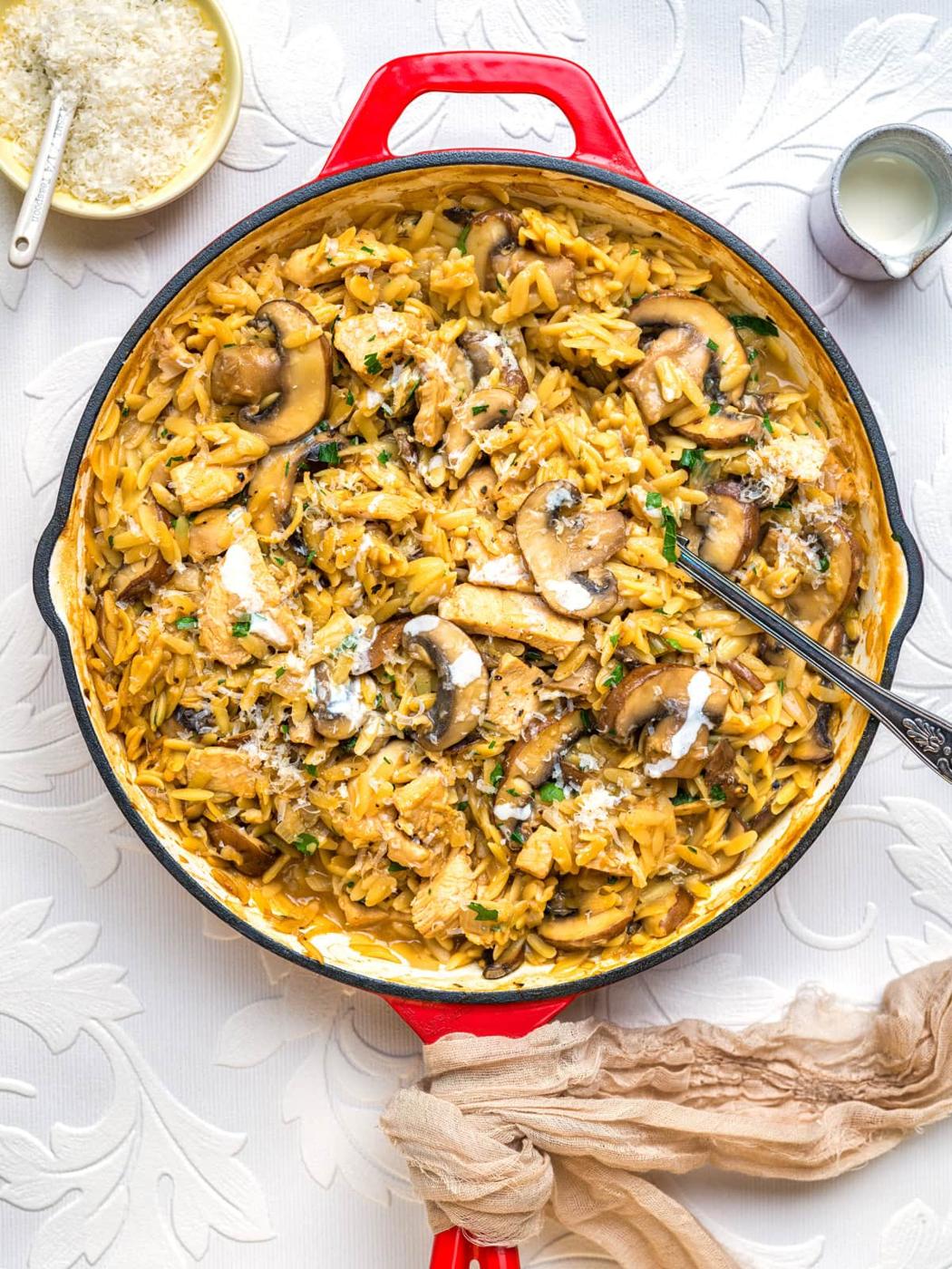 A red skillet filled with creamy one pot marsala orzo, mushrooms, and herbs sits on a white patterned tablecloth. Nearby are a bowl of grated cheese, a small cup of cream, and a beige napkin—perfect for using leftover turkey.
