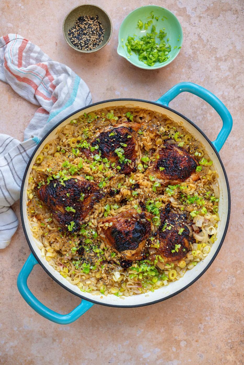 A blue-handled pot filled with browned chicken thighs on top of rice, garnished with chopped green onions and black sesame seeds. Nearby are small bowls of sesame seeds and chopped green onions, and a striped kitchen towel.