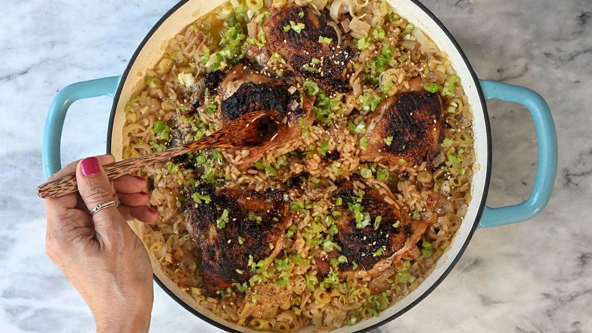 A hand with pink nail polish uses a wooden spoon to serve a dish of browned chicken thighs over rice and vegetables in a round, blue-handled pot on a marble surface.