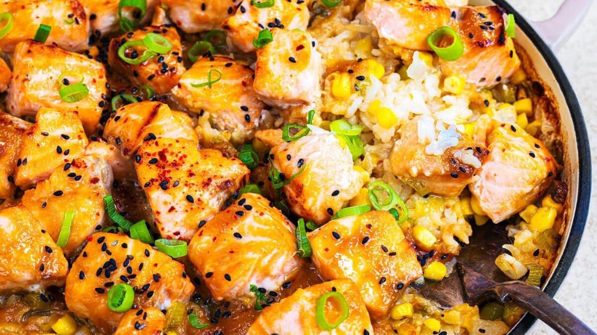 A pan filled with cooked salmon chunks on top of creamy rice with corn, garnished with sliced green onions and black sesame seeds, with a serving spoon on the side.