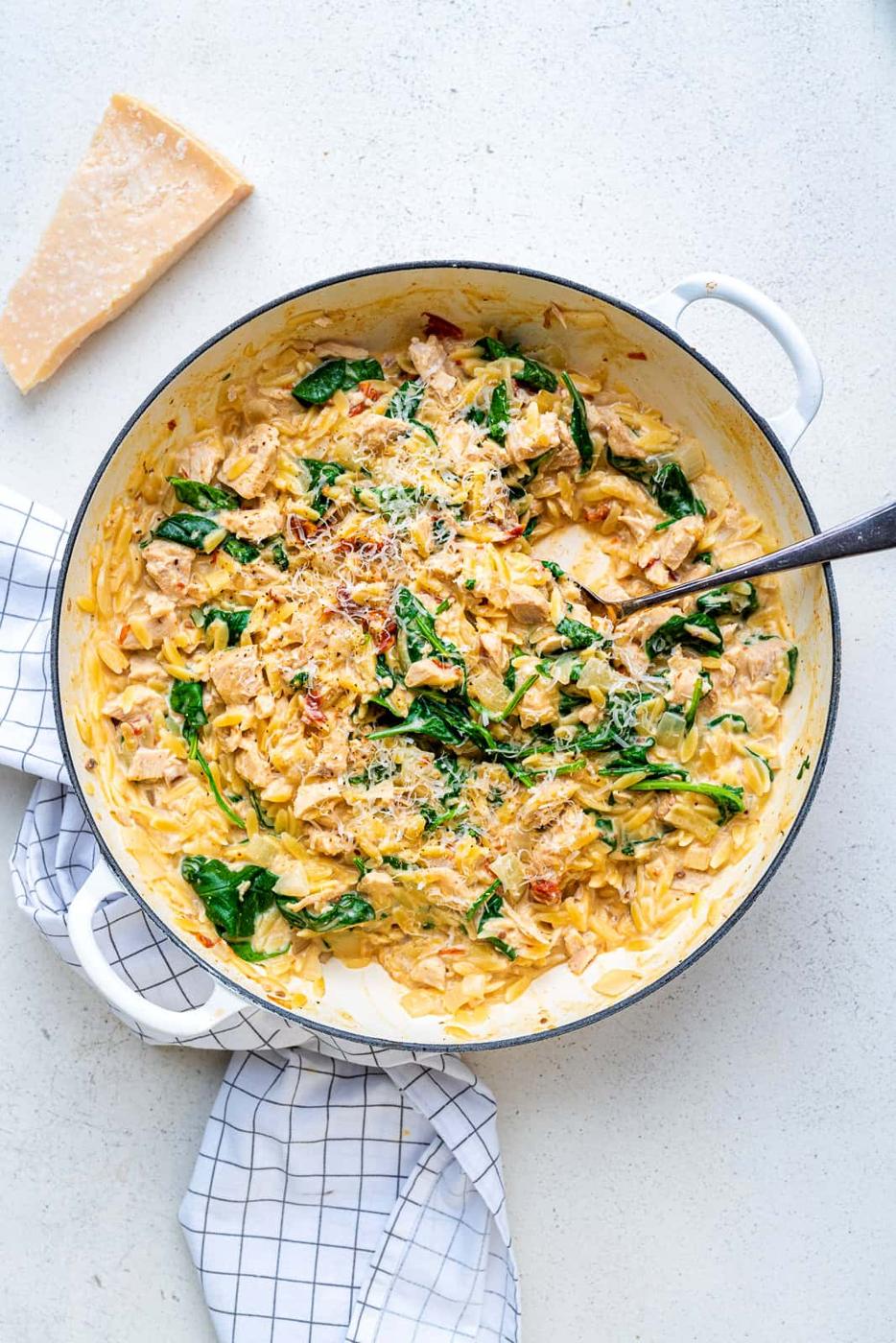 A white skillet filled with creamy One-Pot Tuscan Orzo, spinach, and sun-dried tomatoes, topped with grated cheese. A silver spoon rests in the orzo, and a wedge of parmesan sits nearby on a light countertop with a white towel.