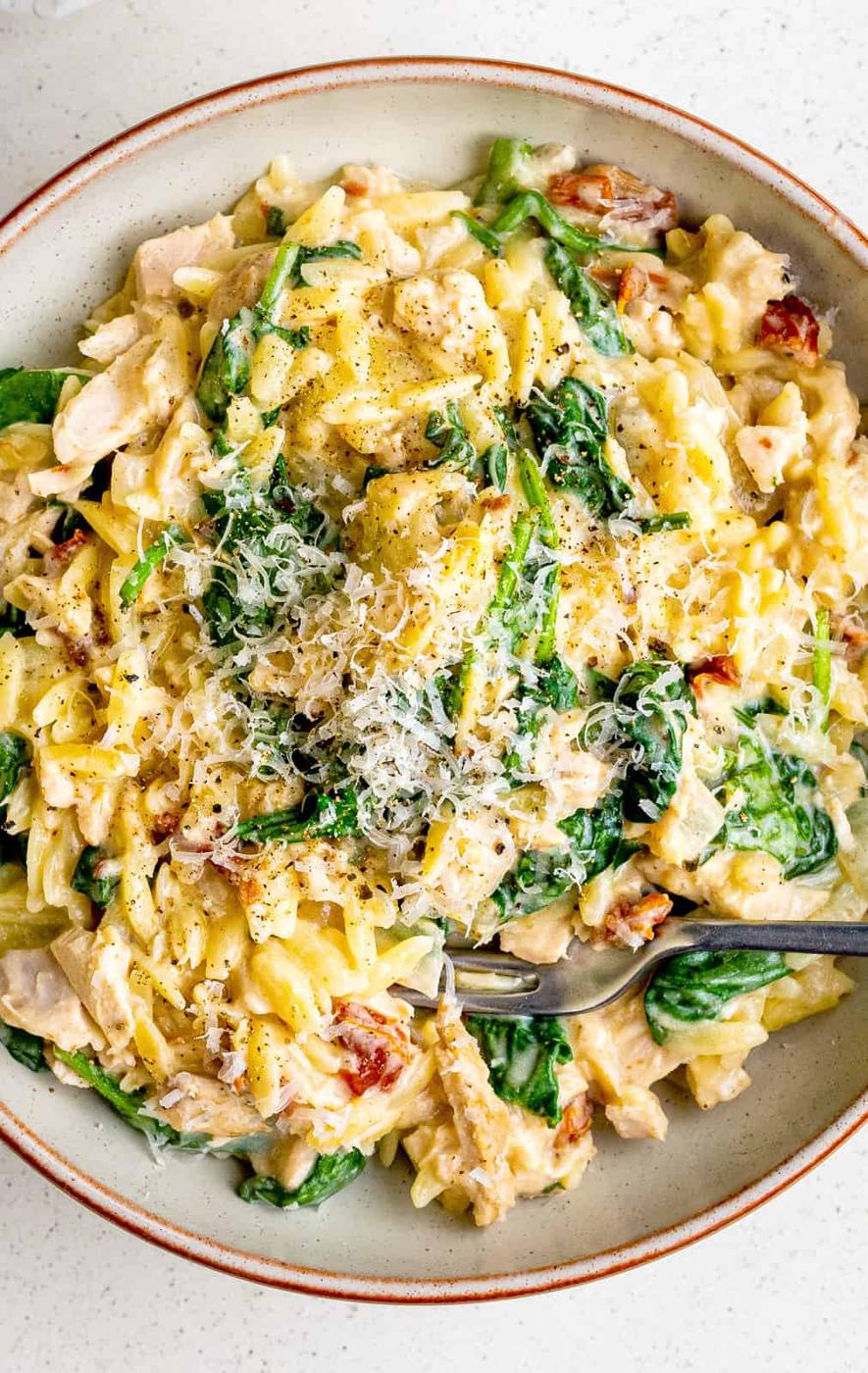 A plate of creamy One-Pot Tuscan Orzo mixed with spinach, sun-dried tomatoes, and shredded chicken, topped with grated cheese and black pepper. A fork and spoon are placed in the bowl, ready to eat.