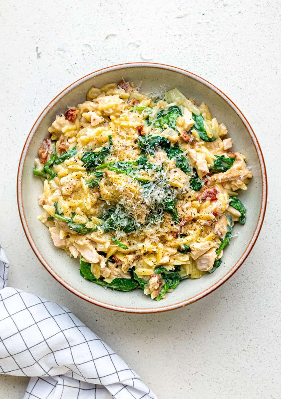 A bowl of creamy One-Pot Tuscan Orzo with spinach, shredded chicken, sun-dried tomatoes, and grated cheese on top sits on a light-colored surface next to a white cloth with a black grid pattern.