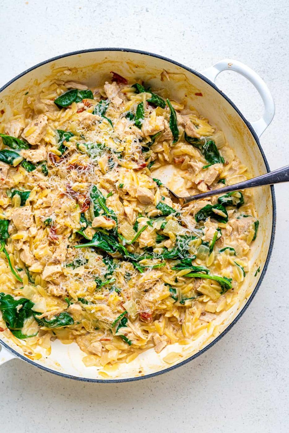 A white pan filled with creamy One-Pot Tuscan Orzo, spinach, shredded chicken, and sun-dried tomatoes, topped with grated cheese. A spoon rests in the pan on a light-colored surface.