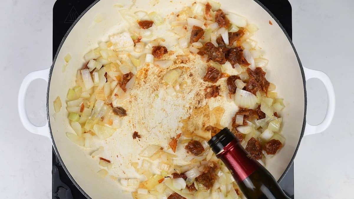 Chopped onions and browned tomato paste are sautéing in a white pan as a bottle pours liquid—likely wine—into the mixture, forming the flavorful base for a One-Pot Tuscan Orzo with leftover turkey on the stovetop.