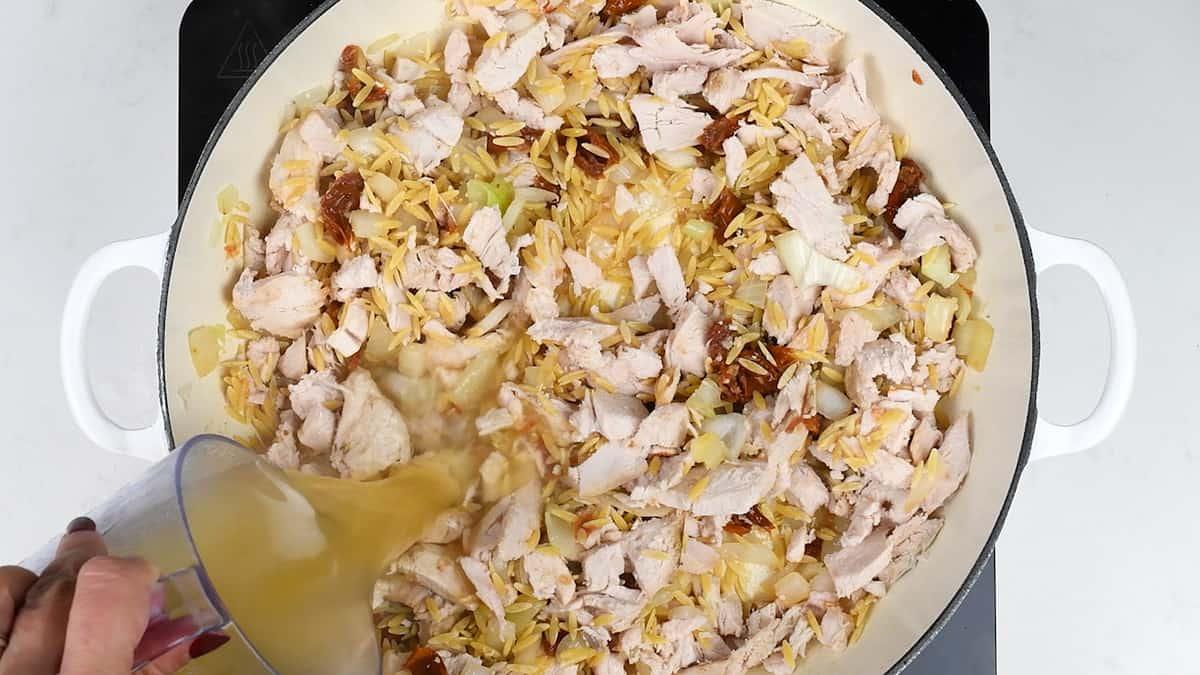 Chopped cooked chicken, onions, and slivered almonds are sautéing in a large white pan as broth is poured in, creating the base for a flavorful One-Pot Tuscan Orzo.