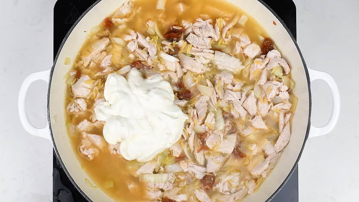A large white pot on a stovetop contains chopped cooked turkey, onions, sun-dried tomatoes, broth, and a dollop of sour cream or yogurt—perfect for making a delicious One-Pot Tuscan Orzo with leftover turkey.