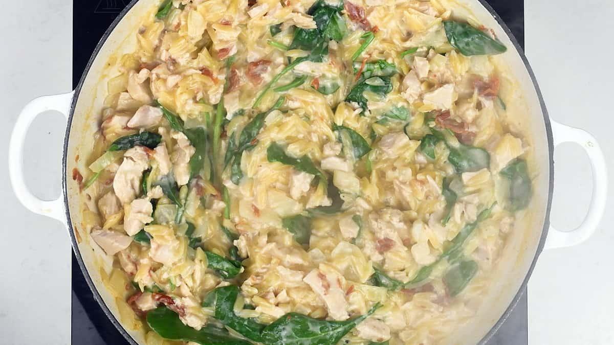 A white pot filled with creamy One-Pot Tuscan Orzo, chicken, cabbage, spinach, and sun-dried tomatoes cooking on a stovetop.