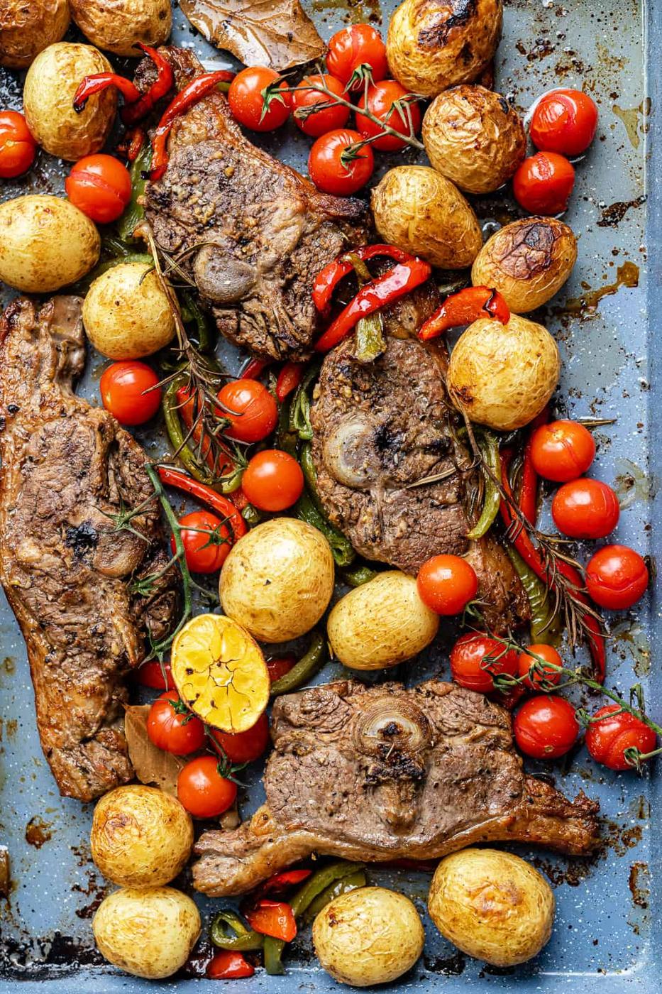 Lamb chops in oven roasted with whole baby potatoes, cherry tomatoes, red peppers, fresh rosemary, and a halved garlic bulb—all golden and slightly charred on a metal baking tray.