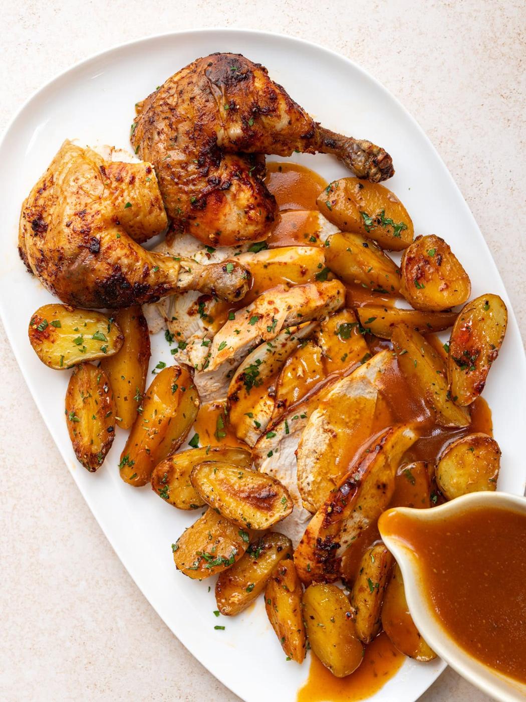 Roast chicken served on a white platter with golden roasted potatoes and brown gravy, garnished with chopped herbs. A small gravy boat sits beside the platter.