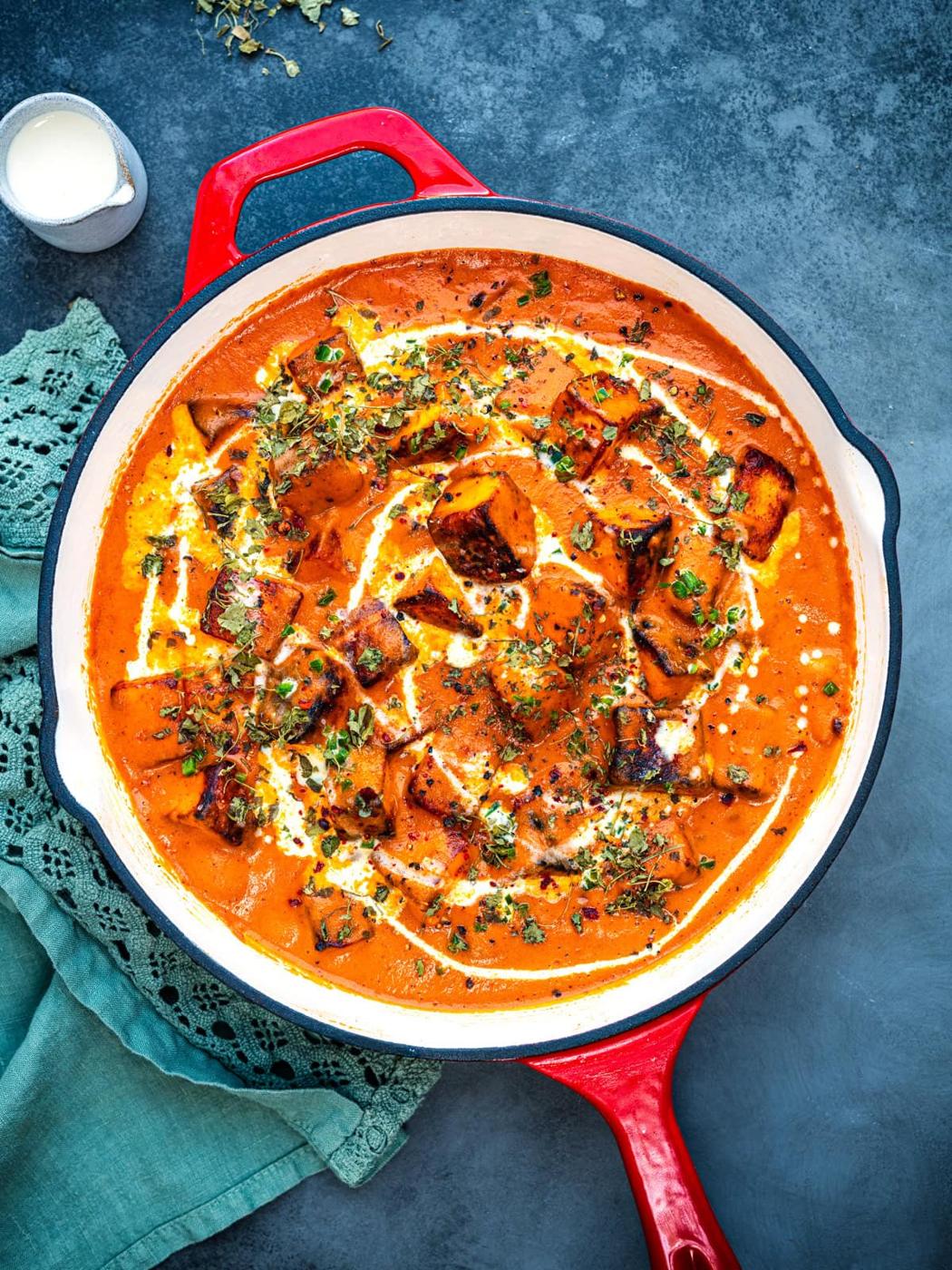 A red pan filled with creamy orange Paneer Butter Masala curry, topped with grilled paneer cubes, drizzled with cream, and garnished with herbs. A green cloth serviette and a small pot of cream sit nearby on a dark surface.