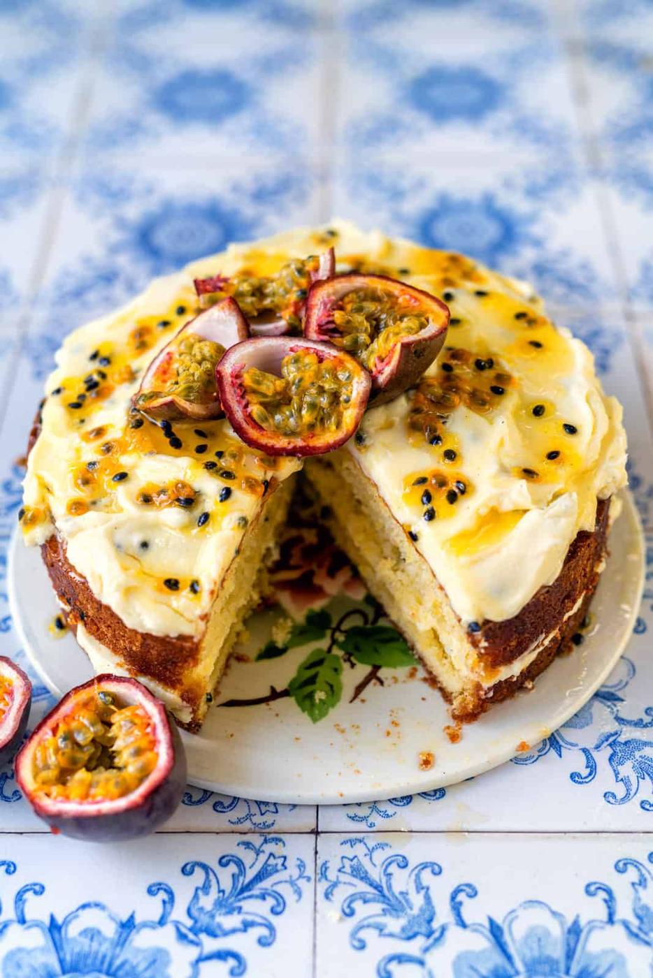 An Easy Passion Fruit Cake with white frosting and passion fruit topping sits on a plate. A slice is missing, revealing the cake’s moist, yellow interior. Halved passion fruits and seeds decorate the cake on a blue patterned surface.