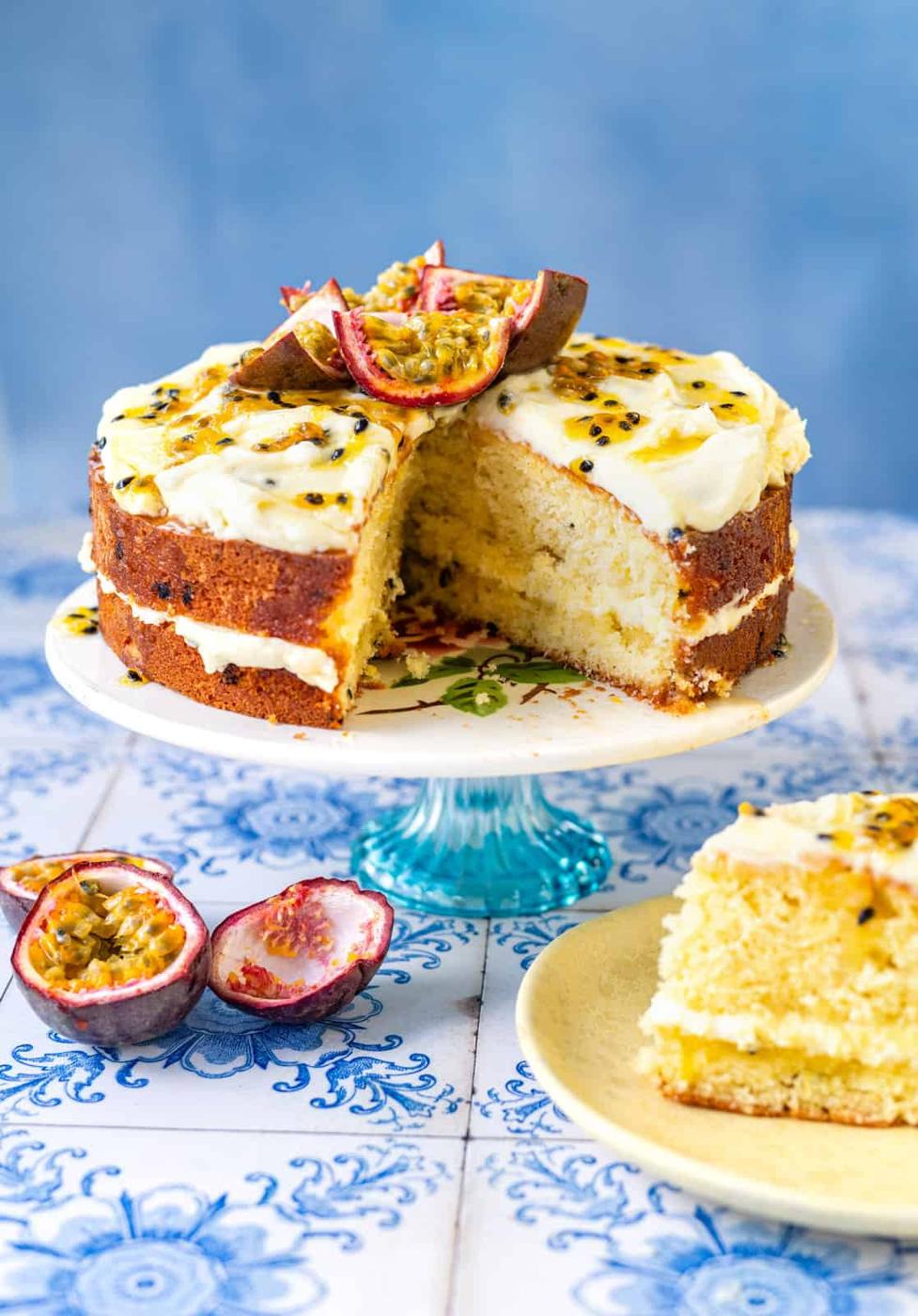 An Easy Passion Fruit Cake with white frosting sits on a blue cake stand. Topped and filled with passionfruit, it has a slice cut out and placed on a plate in front. Fresh passionfruit halves are displayed nearby.