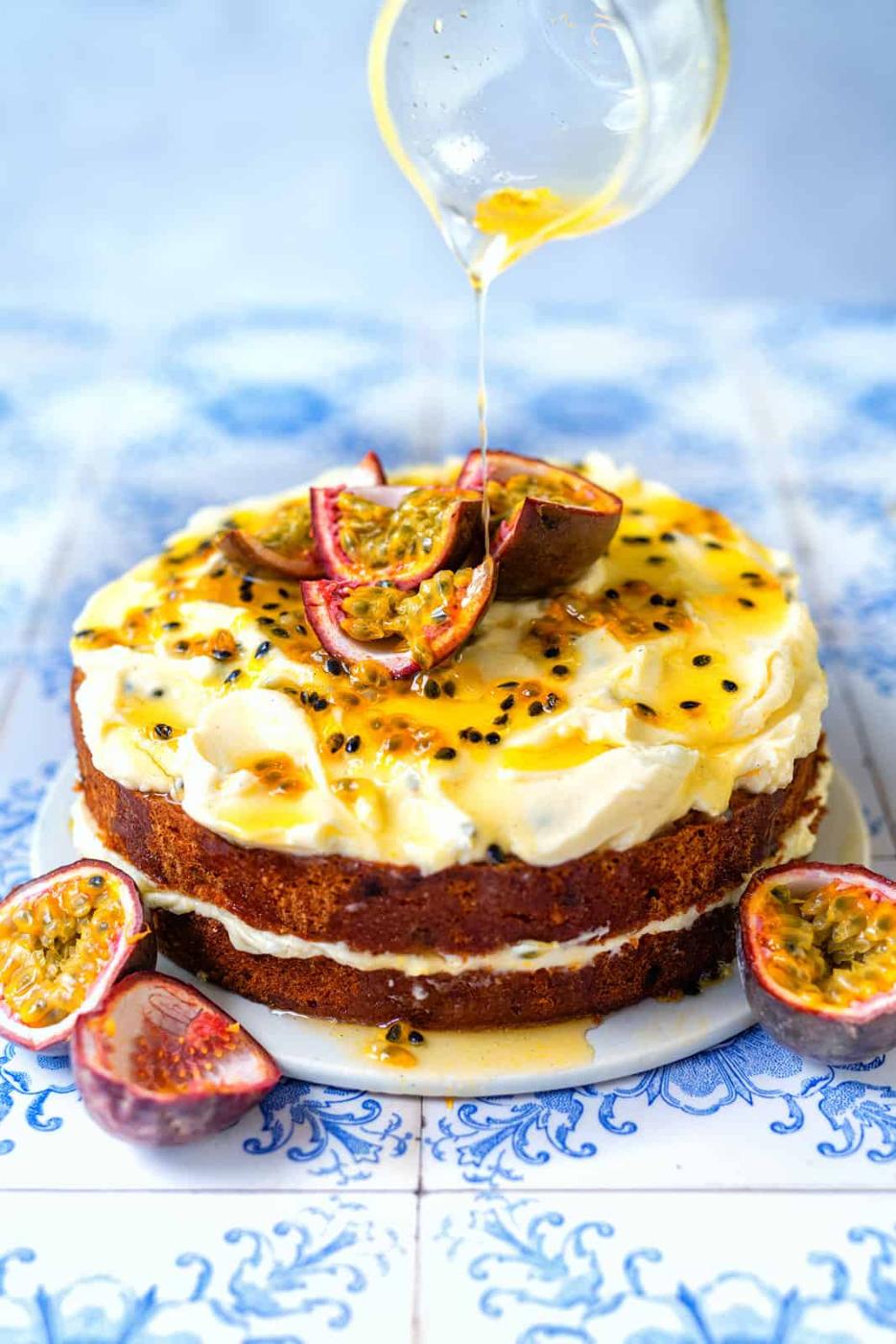 An Easy Passion Fruit Cake with cream frosting and passionfruit topping sits on a blue and white tiled surface. Halved passionfruit decorate the cake as syrup is poured over from a glass container above.