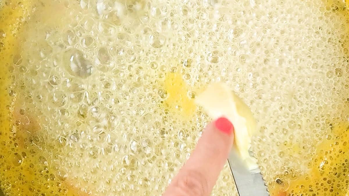 A close-up of a bubbling golden liquid in a pan, with a hand spreading Passion Fruit Jam and butter using a knife, fingernails painted pink.