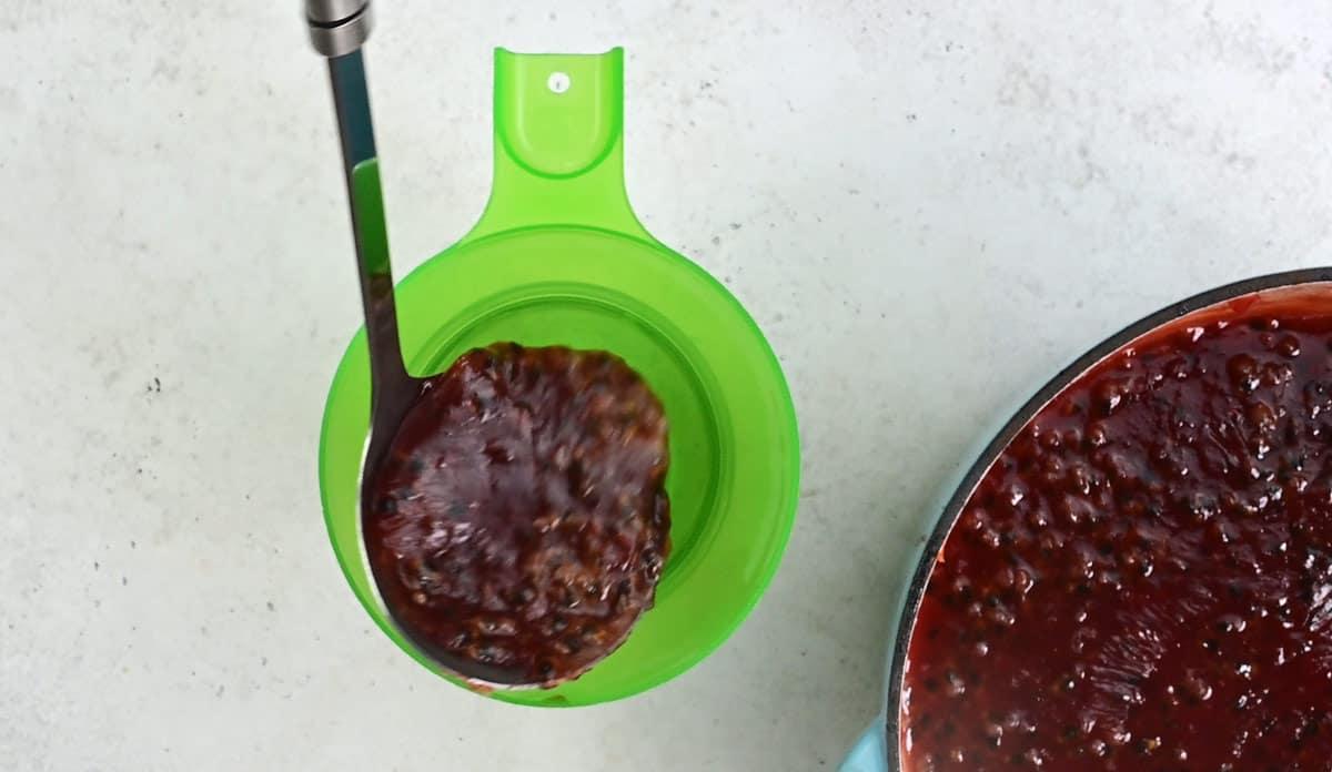 A ladle pours chunky, dark red passion fruit jam through a green funnel into a glass jar, with a pot of jam visible to the right on a light countertop.