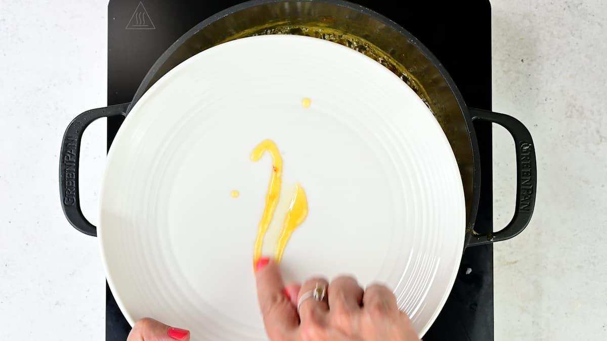 A hand with red nail polish uses a finger to draw a line of Passion Fruit Jam or yellow sauce on a white plate, which is held above a black stovetop with a pot visible underneath.