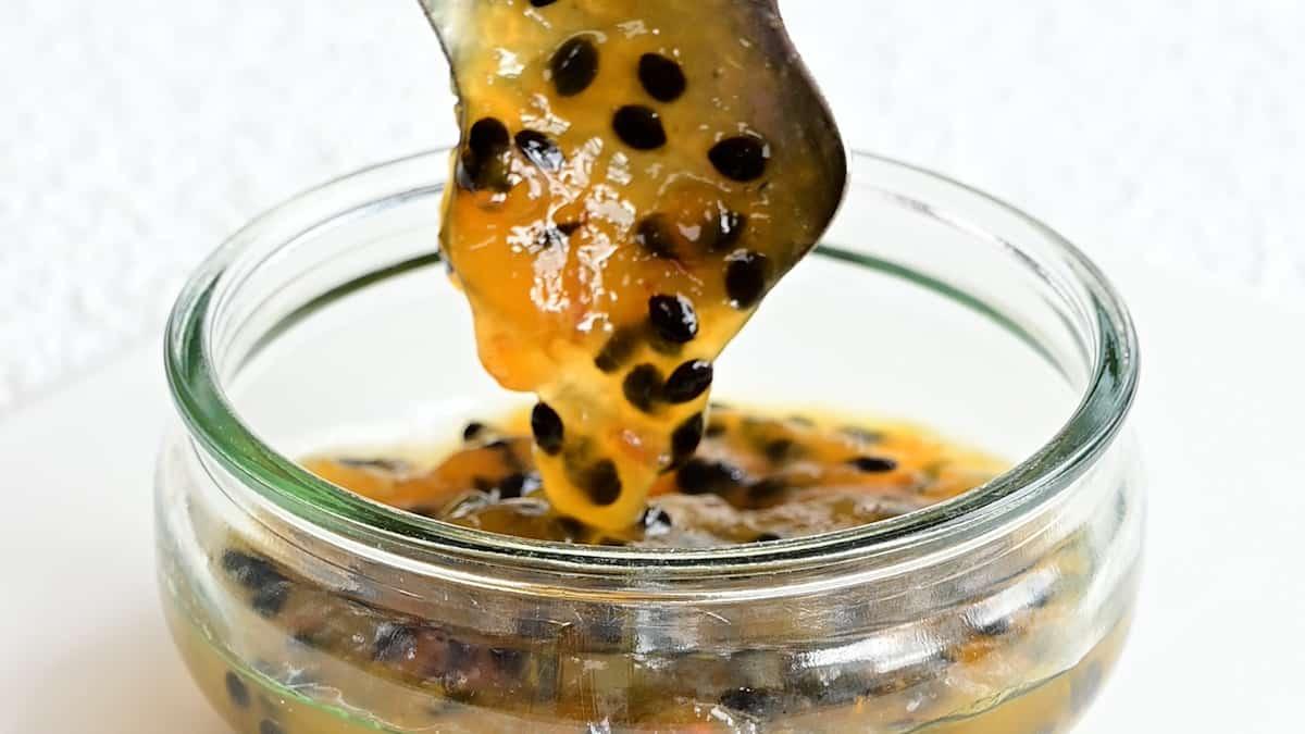 A spoon holds up thick, orange-yellow Passion Fruit Jam with black seeds over a small glass bowl filled with the same vibrant mixture.