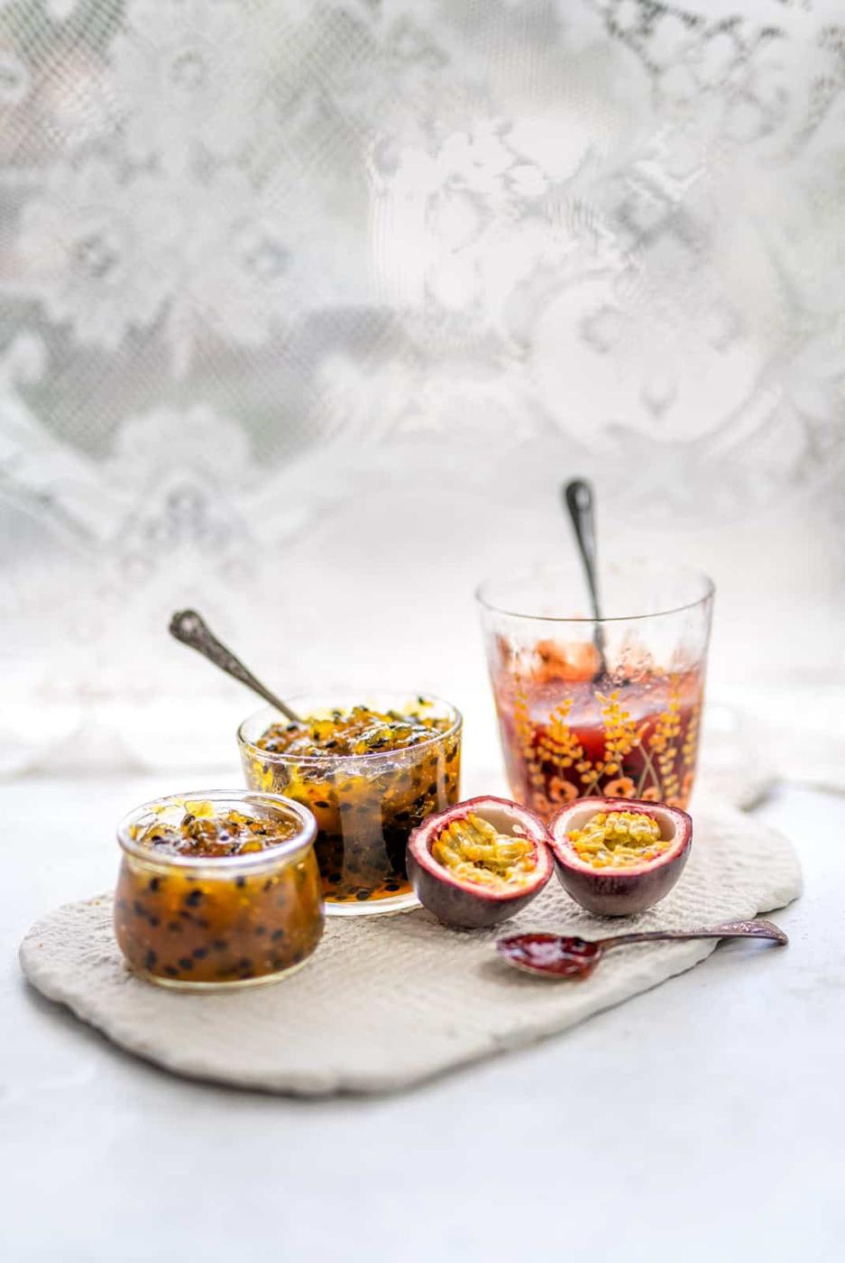 Three small glass jars of Passion Fruit Jam sit on a textured cloth, with two halved passion fruits and a decorative glass of juice nearby. Silver spoons rest in the jars and glass, set against the soft backdrop of a lace curtain.