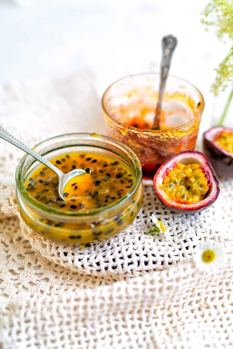 Two glass jars with spoons contain Passion Fruit Jam; one is fuller, the other nearly empty. A halved passion fruit rests nearby, all arranged on a textured white crochet surface.